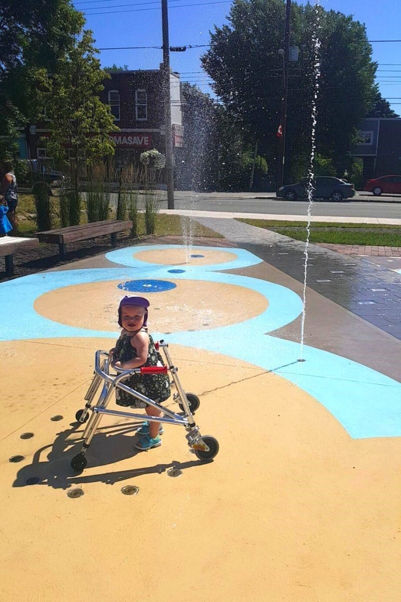A young child using a walker plays on an accessible splash pad