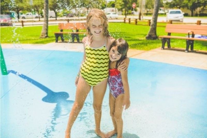 Two girls standing arm in arm on a splash pad, soaked through